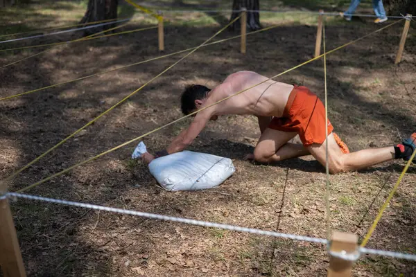 Açık hava engelli parkurunda ağ altında sürünen sporcu, animasyona katılan kum torbası taşıyor. meydan okuma, macera, yarış, fitness, takım çalışması, açık hava