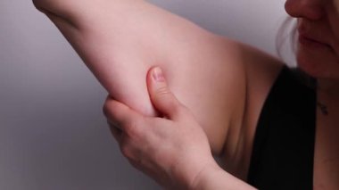 Genç bir kadının kolunun üst kısmında koyu gri bir arkaplanda fazla yağ bulunan deriyi kavrayıp sallarken çekilmiş bir fotoğrafı. Gevşek ve sarkık kasları sıkıyorum. Fazla kilolu konsept. 