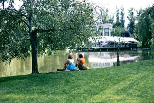 Göl kenarındaki bir parkta yeşil çimlerde oturan iki kız..
