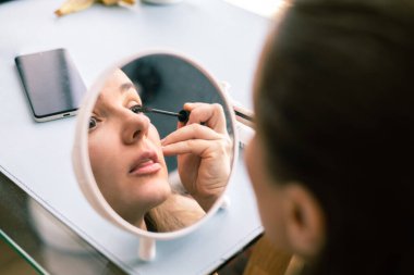 Güzel bir kadın sabah kirpiğinde rimel kullanıyor. Gülümseyen genç bir kadın makyaj yapıyor ve aynaya bakıyor. Güzel kız evde maskara sürüyor..