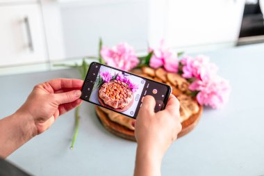 Mutlu pasta şefi kadın rahat bir mutfakta cep telefonuyla meyveli turta fotoğrafı çekiyor.