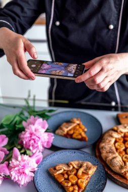 Mutlu pasta şefi kadın rahat bir mutfakta cep telefonuyla meyveli turta fotoğrafı çekiyor.