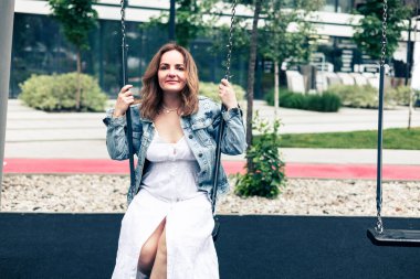 Genç, mutlu, beyaz bir kadın şehirdeki çocuk parkında salıncakta sallanıyor.