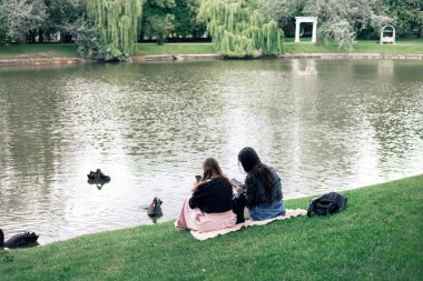 Doğada gölün kenarında otururken telefonu kullanan iki kişi. Sakin bir kırsalda, göl kıyısında dinlenen iki arkadaşın arka görüntüsü