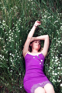 Genç sarışın bir kadın, kır çiçekleriyle çevrili yemyeşil çimlerin üzerinde uzanıyor, doğada huzurlu bir anın tadını çıkarıyor..