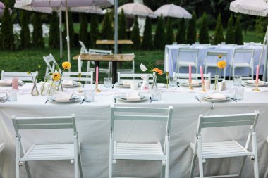 Güneşli bir günde, düğün resepsiyonunda misafirler için dışarıda masalar düzenlenmiş. Beyaz ahşap sandalyeler, düğün töreni için evin dışına kurulmuş..