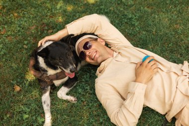 Dostum, rahatla ve çimlerin üzerinde köpekle uzan. Parkta sevgi ve destek için. Manzara, erkek ya da evcil hayvan sahibi ve köpeği doğa dostu.