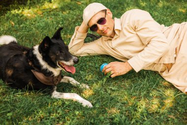 Dostum, rahatla ve çimlerin üzerinde köpekle uzan. Parkta sevgi ve destek için. Manzara, erkek ya da evcil hayvan sahibi ve köpeği doğa dostu.