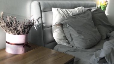 Sabahın erken saatlerinde dağınık bir yatak, güneş ışınları ve kırışık yatakta gölgeler. Yatak örtüsü. Yatağın yanındaki masada bir buket çiçek var. Kapalı beyaz sayfa, yavaş çekim. 