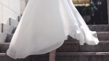 Beyaz gelinlik giymiş genç bir kadın merdivenle aşağı iniyor. Topuklu beyaz ayakkabılar.