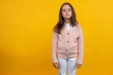 Pembe ceketli ve beyaz pantolonlu genç bir kız sarı arka planda poz veriyor.