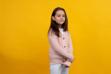 Pembe ceketli ve beyaz pantolonlu genç bir kız sarı arka planda poz veriyor.