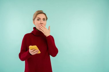 Kırmızı kazaklı, mavi arka planda akıllı telefonu olan, şaşırmış genç bir kadın.