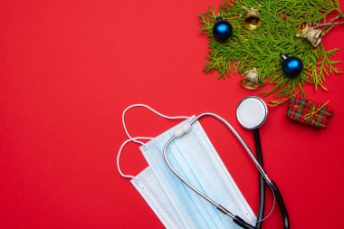 Bayram süsleri, tıbbi steteskop ve kırmızı arka planda yüz maskeleri olan uzun, geniş bir Noel afişi. Sağlık görevlileri için Noel tebrik kartı.