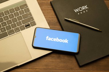 Kyoto Şehri, Japonya - 31 Temmuz 2023: Akıllı telefon ekranındaki Facebook logosu ahşap masa arkasındaki Apple Macbook klavyesine yerleştirildi.