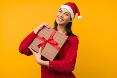 Mutlu, heyecanlı, Noel Baba şapkalı, sarı arka planda hediye kutusu olan genç bir kadın.
