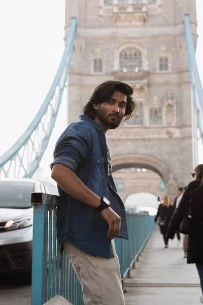 Close up handsome indian stylish guy in London is opposite of London Bridge. He wears a denim jacket and he is looking away from camera. High quality photo