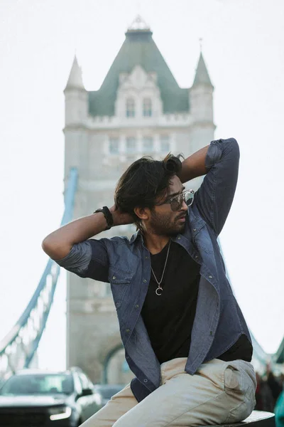 Close up handsome stylish guy in London is opposite of London Bridge and red bus, man siting on Tower Bridge. He wears a denim jacket and he is looking away from camera. High quality photo