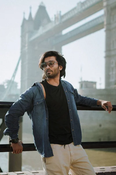 Young indian man standing on Tower Bridge. He wears a denim jacket, sunglasses and he is looking away from camera. High quality photo