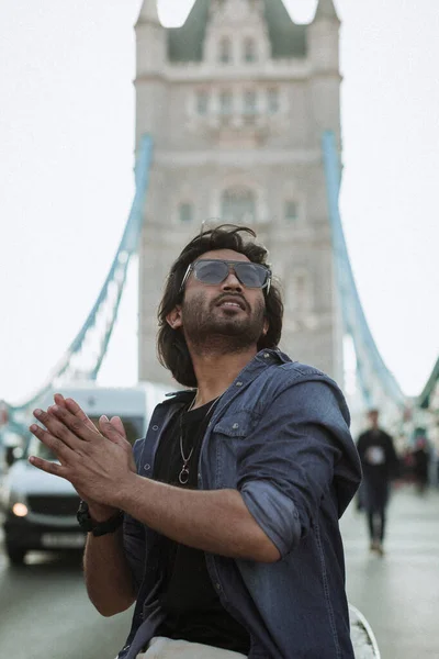 Close up handsome stylish guy in London is opposite of London Bridge, man standing on Tower Bridge. He wears a denim jacket, sunglasses and he is looking away from camera. High quality photo