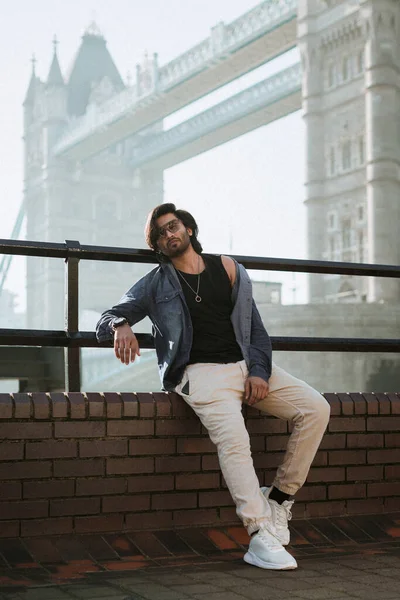 Handsome stylish guy in London is opposite of London Bridge, man standing on Tower Bridge. He wears a denim jacket, sunglasses and he is looking to the camera. High quality photo