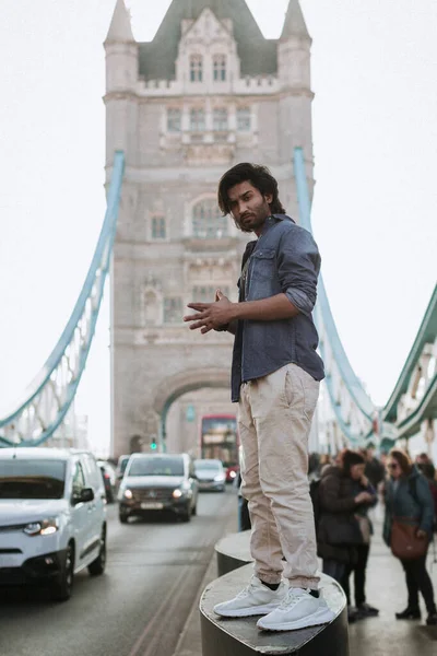Handsome stylish guy in London is opposite of London Bridge, man standing on Tower Bridge. He wears a denim jacket, sunglasses and he is looking away from camera. High quality photo
