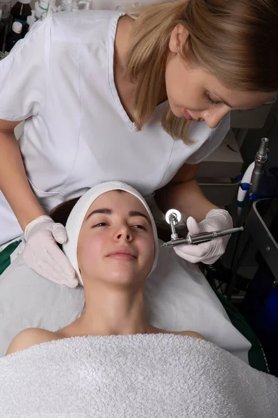 Beautiful girl is receiving anti-aging rejuvenation procedure at spa salon. She is lying with pleasure. Beautician is lifting laser near her cheek. High quality photo