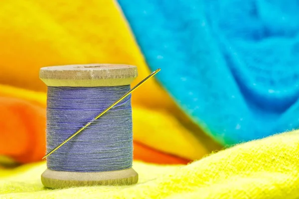 Spool of purple cotton thread and sewing needle sitting on bright colored clothing. Textile tailoring concept with focus on foreground.