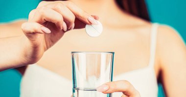 Baş ağrısını dindirmek için uyuşturucu alan bir kadın. Birkaç hap al, bir bardak su tut. Baş ağrısına karşı ilaç alan genç bir kadın..