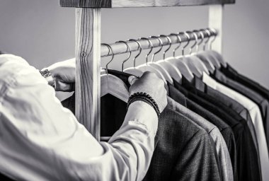 Terzi, terzilik. Şık erkek kıyafeti. Erkek takımları peş peşe asılıyor. Klasik kostüm giymiş moda adamı. Erkek kıyafetleri, butikler. Siyah ve beyaz.
