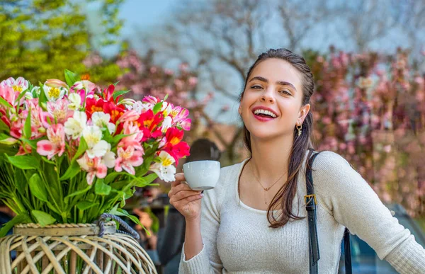 Beautiful woman on a cafe enjoying morning with cup of coffee. She holds a cup, laughing, spring flowers. Woman drink coffee. Spring woman fashion concept. Morning coffee. Spring concept.