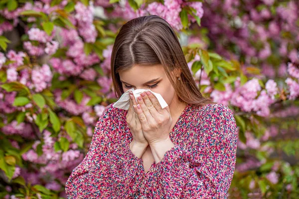 Allergy, sneezing, spring. Woman sneezing in front of blooming tree. Spring allergy concept. Sneezing young girl with nose wiper among blooming trees in park. Pollen allergy, girl sneezing.