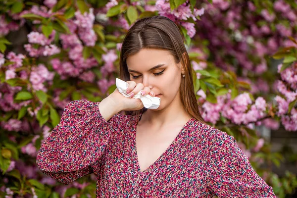 Sneezing young girl with nose wiper among blooming trees in park. Pollen allergy, girl sneezing. Allergy, sneezing, spring. Woman sneezing in front of blooming tree. Spring allergy concept.