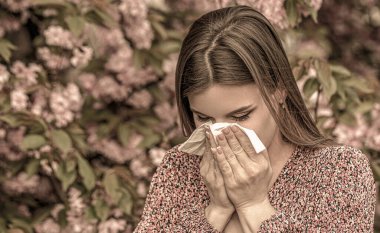 Spring allergy concept. Sneezing young girl with nose wiper among blooming trees in park. Pollen allergy, girl sneezing.