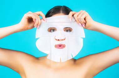 Maskeli güzel bir kadın. Cilt rengiyle ilgilenen bir kız. Cilt bakımı ve güzellik anlayışı. Nemlendirici maske. Mavi arka planda yüzüne çarşaf maskesi takan bir kadın..