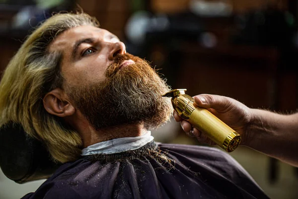 Barber works with a beard clipper. Hipster client getting haircut. Hands of a hairdresser with a beard clipper, closeup. Bearded man in barbershop. Man visiting hairstylist in barbershop.