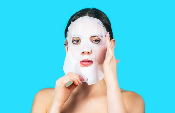 Moisturizing mask. Woman applying sheet mask on her face, on blue background. Beautiful woman with mask, girl taking care of skin complexion. Skin care and beauty concept.