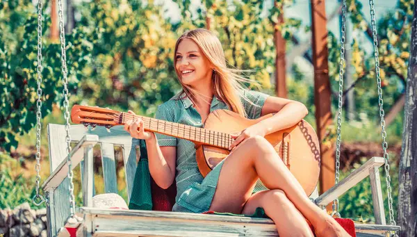 Girl plays guitar in park on a bench, swing. Young woman enjoy playing guitars. Happy hipster woman with guitar. Beautiful young woman plays guitar on park.