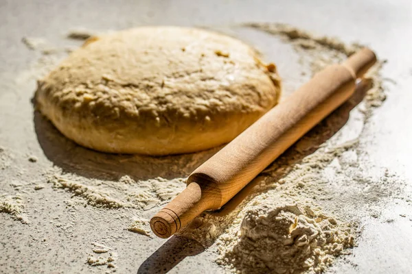 Wooden rolling pin with freshly prepared dough, flour shaker. Fresh raw dough and rolling. Raw dough for pizza or bread baking. Rolled doughs with rolling pin on table covered baking flour.