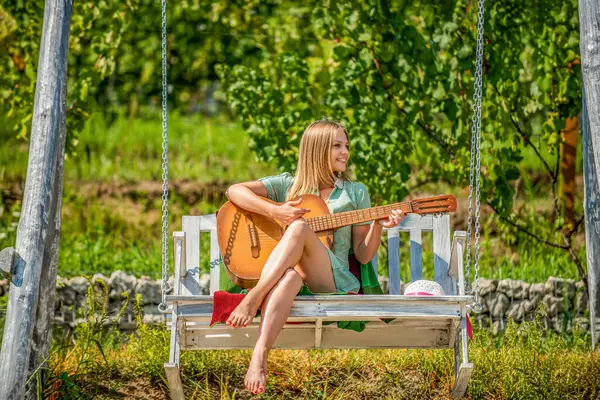 Girl plays guitar in park on a bench, swing. Young woman enjoy playing guitars. Happy hipster woman with guitar. Beautiful young woman plays guitar on park. Happy girl playing guitar outdoor.