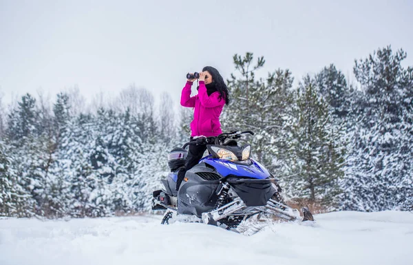 Girl on sports snowmobile in forest. Rides a snowmobile in the mountains. Snowmobile in snow. Woman is riding snowmobile. Travel, girl looks through binoculars. Woman in winter forest with binoculars.