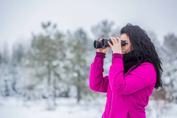 Observation of nature in the winter season. Travel concept, girl looks through binoculars. Woman in winter forest with black binoculars. Hunter woman in the snowy forest looks through binoculars.