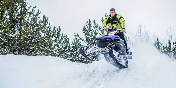 Athlete on a snowmobile moving in the winter forest in the mountains. Man and fast action snowmobile jumping. Jumping on a snowmobile on a background of winter forest. Bright snowmobile.