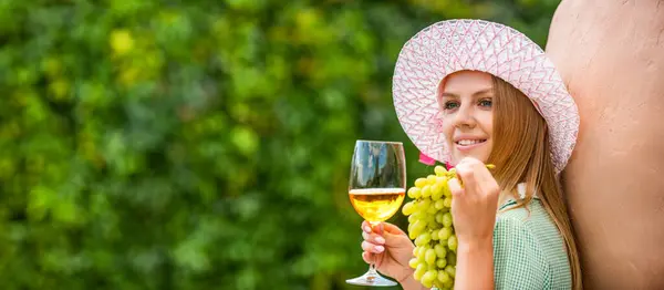 Wine tasting in outdoor winery. Grape and wine making. Woman tasting wine in vineyard. She is showing glass of wine. Woman holding glass of white wine in vineyard, showing grapes.