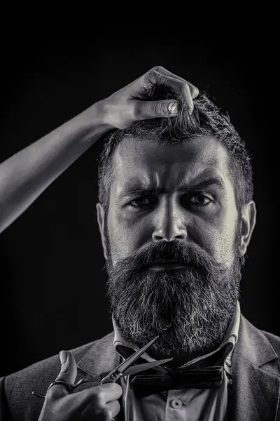 Bearded man hipster barber with beard and moustache in suit holds sharp scissors and cut hair in hairdresser salon. Barber scissors, barber shop. Brutal man, moustache. Black and white.