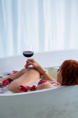 Köpüklü bir küvette şarap ve şampanya içen güzel bir kadın. Güzel seksi kız spa tedavisinde rahatlıyor. Gül yapraklarıyla banyo yaparken elinde bir bardak şarap tutan bir kadın. Koca memeli, seksi kız..