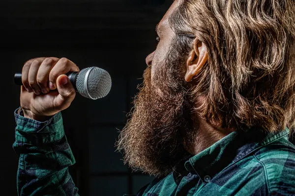 Guy with beard singing in microphone. Bearded man wearing checkered shirt. Male karaoke singer. Bearded man singing with microphone. Man with a beard holding a microphone and singing.