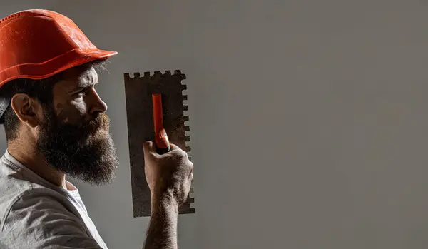 Plasterer renovating wall. Profile of bearded builder. Worker with plastering tools. Renovating house concept. Builder man in safety helmet. Tool, trowel, handyman, man builder. Mason tools, builder.