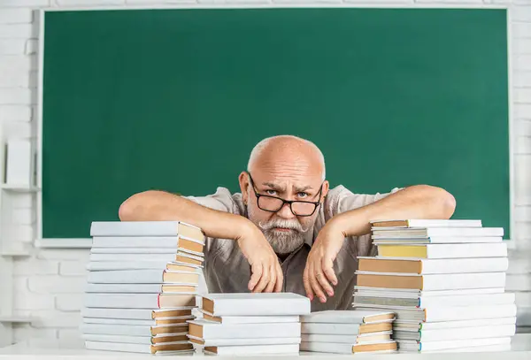 Teacher with too many books to read before exam. Teacher with books in school. Man school teacher standing in classroom. Old man teachers on green board. Funny teacher hold many books at school.