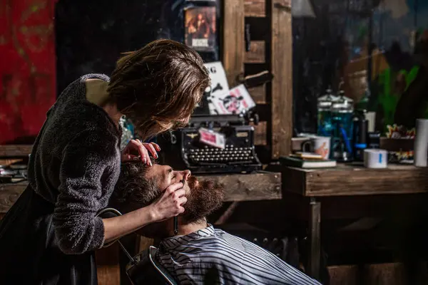 Barber girl shaves his beard with a dangerous razor. Women barber shaves his beard. Barber woman shaving a bearded man in a barber shop.